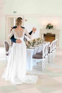 Ein Paar tanzt in einem eleganten Raum mit einem langen, mit Blumenarrangements geschmückten Esstisch und fängt damit die Essenz der Hochzeitsplanung ein. Die Frau trägt ein weißes Spitzenbrautkleid und der Mann einen dunklen Anzug, während im Hintergrund ein dekorativer Kamin mit Kerzen romantisch flackert.