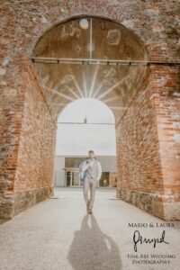 Ein Mann im hellen Anzug schreitet selbstbewusst durch einen sonnenbeschienenen Backsteinbogen, während goldene Strahlen seinen Weg erhellen. Die Stadtarchitektur schafft die Kulisse für diesen Moment der Eleganz und Verheißung. Das Bild, perfekt als Inspiration für Hochzeitsplanung, ist in der unteren rechten Ecke mit dem Namen eines Fotogeschäfts gekennzeichnet.