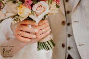Nahaufnahme einer Braut und eines Bräutigams während ihrer Hochzeit, die einen wunderschön arrangierten Brautstrauß halten. Die mit einem Ehering geschmückten Hände der Braut umfassen sanft Rosen und Grünpflanzen. Der Bräutigam in einem hellen Anzug steht liebevoll neben ihr.