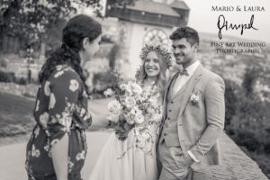 Ein Schwarzweißfoto fängt die Hochzeit wunderschön ein und zeigt Braut und Bräutigam, die im Freien mit einer Frau posieren. Die mit Blumen gekrönte Braut hält einen Blumenstrauß, während der Bräutigam eine Blumenboutonniere trägt. Hinter ihnen steht ein Gebäude mit einer Uhr – eine perfekte Momentaufnahme eines eleganten Heiratsmoments.