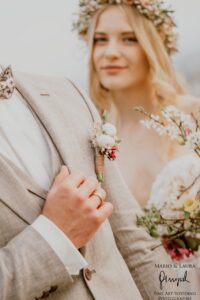 Eine Nahaufnahme von Braut und Bräutigam während ihrer Hochzeit. Der Bräutigam, in einem hellen Anzug mit einer Boutonniere, hält sanft die Hand der Braut. Hinter ihm trägt die Braut eine Blumenkrone und umklammert ihren Blumenstrauß, was die Liebe und Freude des Heiratens verkörpert.