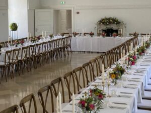 Eleganter Bankettsaal mit zwei langen, U-förmig angeordneten Esstischen, perfekt für eine Hochzeit, geschmückt mit Blumengestecken und hohen Kerzen. Die Tische sind mit Holzstühlen gesäumt und im Hintergrund ist ein dekorativer Kamin mit weiteren Blumenarrangements zu sehen. Ideal für unvergessliche Hochzeitsfeiern.