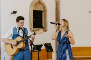 Ein Mann in einer blauen Weste spielt Akustikgitarre, während eine Frau in einem blau gestreiften Kleid in ein Mikrofon singt und so die perfekte Atmosphäre für eine intime Hochzeit schafft. Sie treten drinnen auf, eingerahmt von einer Steinmauer mit einem kleinen Bogenfenster, vor ihnen stehen Notenständer.