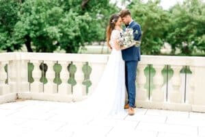 Braut und Bräutigam im Hochzeitskleid stehen auf einer Steinterrasse mit üppigen grünen Bäumen im Hintergrund. Während sie ihre Hochzeit feiern, hält die Braut einen Blumenstrauß in der Hand, während der Bräutigam einen blauen Anzug mit braunen Schuhen trägt. Sie umarmen sich freudig und fangen einen perfekten Moment ihrer Hochzeitsreise ein.