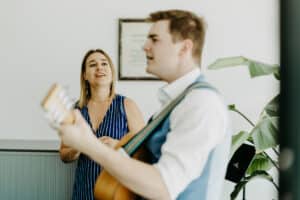 Bei dieser entspannten Hochzeitsfeier im Innenbereich singt eine Frau im blauen Kleid fröhlich, während ein Mann in einer Weste Gitarre spielt. Eine Topfpflanze trägt zur bezaubernden Atmosphäre bei.
