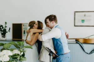 Zwei Personen in formeller Kleidung umarmen sich, umgeben von einem anderen Paar, das sich ebenfalls umarmt und die Freude der Hochzeit einfängt. Eine Gitarre und gerahmte Urkunden hängen an der Wand und verleihen dem Raum Charme. Eine Blattpflanze schmückt den Raum und bietet eine ruhige Kulisse für diesen Moment der Hochzeitsplanung.