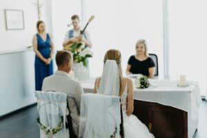 Bei einer wunderschönen, hellen Hochzeitszeremonie mit weißem Dekor sitzen Braut und Bräutigam an einem Tisch und begeben sich auf ihre Hochzeitsreise. Eine Frau leitet die Zeremonie, ein Mann mit einer Gitarre sorgt für musikalischen Beifall, und eine weitere Frau beobachtet die Zeremonie im Hintergrund.