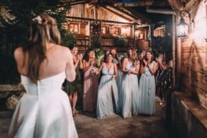Brautjungfern in hellblauen Kleidern und andere Gäste reagieren freudig, als sie einer Braut im weißen Kleid gegenüberstehen. Der Hochzeitsempfang findet in einem rustikalen Holzsaal mit sanfter Beleuchtung statt, was eine warme und festliche Atmosphäre schafft.