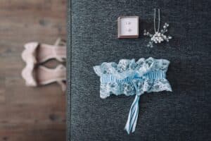 Ein Paar Perlenohrringe in einer Schachtel, ein zartes Haar-Accessoire mit Perlen, ein blaues Strumpfband aus Spitze und rosa Schuhe mit hohen Absätzen sind sorgfältig auf einer grauen gepolsterten Oberfläche und einem Holzboden arrangiert – perfekt für eine Hochzeitsfeier.
