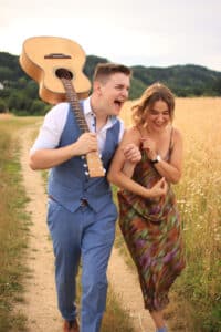 Ein Mann im blauen Anzug lacht und trägt eine Gitarre auf der Schulter, während er mit einer Frau im bunten Kleid einen Feldweg entlanggeht. Sie wirken fröhlich und halten sich vor Feldern und Hügeln die Arme – vielleicht stellen sie sich ihre zukünftige Hochzeit inmitten dieser malerischen Landschaft vor.