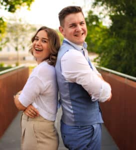Ein lächelnder Mann und eine lächelnde Frau stehen Rücken an Rücken im Freien und verkörpern die Freude am Heiraten. Die Frau trägt ein weißes Hemd und beige Hosen; der Mann trägt eine blaue Weste mit Hosen und ein weißes Hemd. Sie stehen auf einem Gehweg, umgeben von Bäumen und Grün, und genießen ihre gemeinsame Reise in die Hochzeitsplanung.