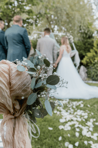 Eine bezaubernde Hochzeitszeremonie im Freien zeichnet sich durch ein Blumenarrangement aus Eukalyptus und weißen Blumen aus, die mit beigem Stoff zusammengebunden sind. Im Hintergrund steht ein Paar zusammen unter einem Bogen, umgeben von Gästen auf einer Rasenfläche – eine perfekte Kulisse für alle, die Wert auf eine sorgfältige Hochzeitsplanung legen.