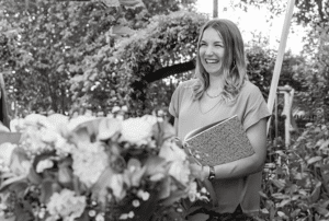 Eine lächelnde Frau steht mit einem Notizbuch in der Hand in einem Garten, umgeben von üppigem Laub und Blumen. Die Szene fängt die freudige Essenz der Hochzeitsplanung inmitten der Schönheit der Natur ein.