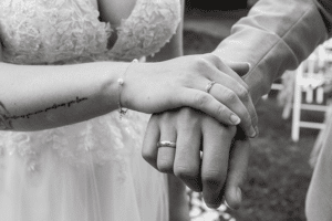 Nahaufnahme der Hände eines Paares, elegant übereinandergelegt, die Eheringe zur Schau stellend. Die Braut in ihrem Spitzenkleid und Armband, der Bräutigam in seinem Anzug. In Schwarzweiß festgehalten, fängt dieser intime Moment die Essenz des Heiratens ein und hebt die komplizierten Details ihrer vereinten Hände hervor.