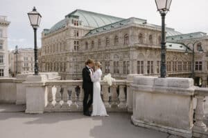 Ein Paar im Hochzeitskleid umarmt sich auf einem Steinbalkon, eingerahmt von zwei Straßenlaternen, die für eine romantische Atmosphäre sorgen. Das historische Gebäude mit einem grünen Dach im Hintergrund bildet die perfekte Kulisse für ihre Hochzeit und fängt einen Moment zeitloser Liebe und eleganter Hochzeitsplanung ein.