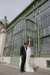 Das Paar feiert seine Hochzeit mit einem Kuss vor einem großen, kunstvoll verzierten Gewächshaus. In formeller Kleidung umklammert die Braut ihren weißen Blumenstrauß, während sie auf einem Steinpflaster unter einem bewölkten Himmel stehen, was ihrem Hochzeitsmoment eine bezaubernde Note verleiht.