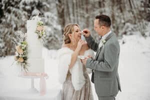 Vor einer winterlichen Kulisse aus Bäumen und Schnee füttert sich ein Paar in Abendgarderobe freudig mit Kuchen neben einer großen, weißen Hochzeitstorte, die mit Blumen geschmückt ist. Die verschneite Umgebung im Freien macht ihren besonderen Moment, in dem sie ihre Verbindung feiern, noch magischer.
