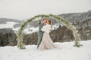 Ein Paar umarmt und küsst sich im Schnee unter einem Blumenbogen und verkörpert eine traumhafte Hochzeit. Die Braut, in einen Pelzschal über ihrem Kleid gehüllt, und der Bräutigam in einem leichten Anzug schwelgen in ihrem Winterwunderland, perfekt für diejenigen, die tief in die Hochzeitsplanung vertieft sind, inmitten ruhiger schneebedeckter Hügel und Bäume.