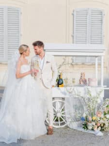 Braut und Bräutigam in eleganter Hochzeitskleidung stoßen mit Champagner vor einem dekorierten weißen Wagen mit Gebäck und Blumen an. Der Hintergrund mit hell gestrichenen Fensterläden und einer hellen Wand verstärkt die romantische Atmosphäre und ist ein Beispiel für eine perfekte Hochzeitsplanung.