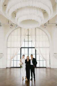 Drei Personen stehen zusammen in einem geräumigen, elegant eingerichteten Raum mit hohen Decken und großen Fenstern, die mit transparenten Vorhängen verhangen sind. Die Person in der Mitte, wahrscheinlich diejenige, die bald heiraten wird, hält einen Blumenstrauß. Sie sind alle in formelle Kleidung gekleidet und lächeln in die Kamera, als würden sie diesen besonderen Hochzeitsmoment genießen.