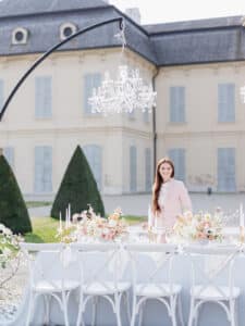 Eine Frau im rosafarbenen Kostüm steht an einem elegant gedeckten Tisch im Freien, der mit pastellfarbenen Blumen und weißen Kerzen geschmückt ist – ideal für eine Traumhochzeit. Die Szene besteht aus weißen Stühlen, einem Kristallkronleuchter und einem prachtvollen Gebäude im Hintergrund, das anspruchsvolle Hochzeitsplanung verkörpert.