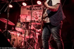Ein Schlagzeuger spielt energisch ein Schlagzeug auf der Bühne, eingerahmt von rotem Licht und einem Hintergrund mit Text. Ein weiterer Musiker ist im Vordergrund teilweise sichtbar und spielt eine Gitarre. Die Atmosphäre ist dynamisch und lebendig.