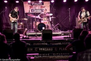 Drei Musiker treten auf einer schwach beleuchteten Bühne unter violettem Licht auf. Einer spielt Gitarre, ein anderer Schlagzeug und der dritte Bass. Hinter ihnen hängt ein Banner mit der Aufschrift „Fourty Finger Circus“. Das Publikum schaut vom Vordergrund aus zu.