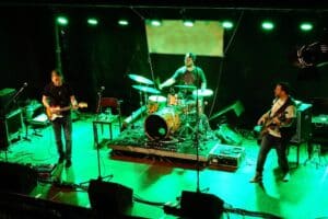 Eine Band tritt auf der Bühne unter grünem Licht auf. Der Gitarrist links spielt eine E-Gitarre, der Schlagzeuger steht mittig hinter einem Schlagzeug und der Bassist rechts spielt eine Bassgitarre. Um sie herum sind Mikrofone und Verstärker aufgestellt.