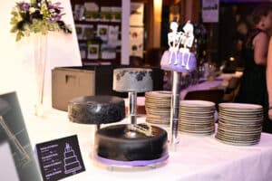 Eine Hochzeitstorte mit drei schwarzen und violetten Torten. Auf der höchsten Torte sitzt eine kleine Skelettfigur eines Paares. In der Nähe sind Teller gestapelt und im Hintergrund ist ein Blumenarrangement zu sehen.