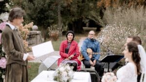Eine Hochzeitszeremonie im Freien mit einer Trauungsleiterin, einem Musiker mit Gitarre und zwei sitzenden Gästen. Die Szene ist von üppigem Grün und rosa Blumen umgeben, was eine romantische Atmosphäre schafft.