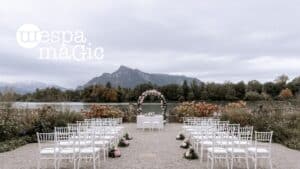 Hochzeitsaufbau im Freien an einem See mit einem mit Blumen geschmückten Bogen an der Vorderseite. Reihen weißer Stühle stehen vor dem Bogen, im Hintergrund sind Berge und bewölkter Himmel zu sehen. Zur Dekoration gehören rosa und weiße Blumen.