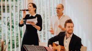 Eine Frau in einem schwarzen Kleid singt in ein Mikrofon und hält Papiere in der Hand. Ein Mann mit Brille klatscht neben ihr. Ein anderer Mann im Anzug spielt Akustikgitarre. Sie befinden sich in einem Gebäude mit großen Fenstern im Hintergrund.