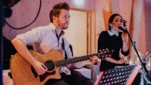 Ein Mann spielt im Sitzen eine Akustikgitarre, neben ihm singt eine Frau in ein Mikrofon. Sie befinden sich in einem warm beleuchteten Raum mit Holzwänden. Vor dem Mann steht ein Notenständer mit Notenblättern.
