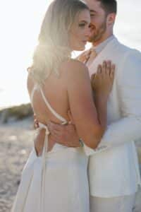 Ein Paar umarmt sich bei Sonnenuntergang am Strand. Die Frau trägt ein rückenfreies weißes Kleid, während der Mann einen hellen Anzug trägt. Sie stehen eng beieinander, während die Sonne einen warmen Schein um sie herum erzeugt. Im Hintergrund sind weicher Sand und eine verschwommene Landschaft zu sehen.