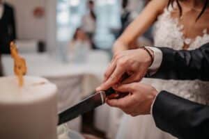 Braut und Bräutigam schneiden gemeinsam ihre Hochzeitstorte an und halten dabei das Messer mit verschränkten Händen. Die Braut trägt ein weißes Spitzenkleid und der Bräutigam einen dunklen Anzug. Die Torte ist elegant dekoriert und mit einer Figur gekrönt.