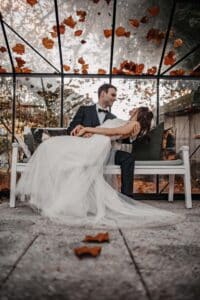 Eine Braut im weißen Kleid und ein Bräutigam im schwarzen Anzug sitzen zusammen auf einer weißen Bank in einem Gebäude mit Glaswänden. Herbstblätter liegen verstreut auf dem Boden und um sie herum fallen weitere Blätter herab. Das Paar blickt sich liebevoll in die Augen.