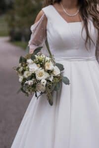Eine Braut in einem weißen Kleid hält einen Strauß weißer und cremefarbener Blumen mit Grünpflanzen in der Hand. Das Kleid hat durchsichtige, gepunktete lange Ärmel. Eine Perlenkette schmückt ihren Hals. Sie steht auf einem gepflasterten Weg mit Grünpflanzen im Hintergrund.