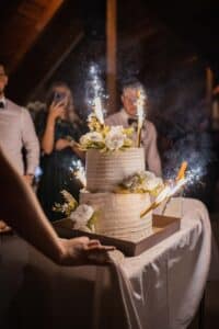 Eine zweistöckige weiße Torte, geschmückt mit weißen Blumen und Wunderkerzen, steht bei einer Veranstaltung auf einem Tisch. Im Hintergrund stehen mehrere Personen, eine hält ein Smartphone, um ein Foto zu machen. Rauch der Wunderkerzen schwebt über der Torte.