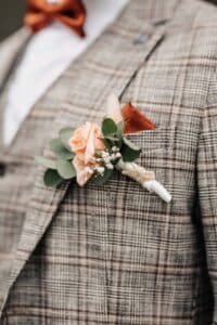 Nahaufnahme einer Person, die einen karierten Anzug trägt und an dessen Revers eine pfirsichfarbene Ansteckblume mit Rosen befestigt ist. Die Ansteckblume ist mit Grünpflanzen und kleinen weißen Blumen verziert und harmoniert mit einem orangefarbenen Einstecktuch in der Tasche.