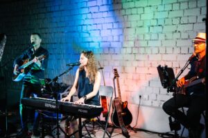 Ein Trio von Musikern tritt auf der Bühne auf: eine Frau spielt Keyboard, ein Mann mit Gitarre und ein weiterer Mann mit Bassgitarre. Sie sind von bunten Bühnenlichtern vor einer weißen Backsteinwand umgeben.