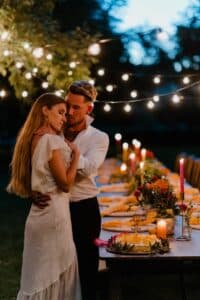 Ein Paar umarmt sich romantisch an einem mit Kerzen und Blumen geschmückten Esstisch im Freien. Darüber hängen Lichterketten und beleuchten die abendliche Kulisse. Die Frau trägt ein weißes Kleid und der Mann ein weißes Hemd.