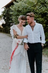 Ein Paar geht einen Kiesweg entlang, hält sich an den Händen und lächelt sich herzlich an. Die Frau trägt ein weißes Kleid mit Blumen, der Mann ein weißes Hemd und eine schwarze Hose, umgeben von üppigem Grün.