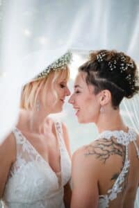 Zwei Frauen in Brautkleidern stehen dicht beieinander und lächeln unter einem transparenten Schleier. Beide tragen Blumenschmuck im Haar. Die eine hat kurze blonde Haare, die andere kurze dunkle Haare und eine Tätowierung auf der Schulter. Die Szene ist intim und fröhlich.