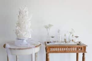 Elegante Hochzeitsdekoration mit einer großen, weißen, dekorativen Torte auf einem runden, mit Tüll bedeckten Tisch. In der Nähe steht ein Holztisch mit Wasserflaschen, zwei Gläsern mit dunkler Flüssigkeit und Zweigen weißer Blumen in Glasgefäßen vor einem schlichten Hintergrund.