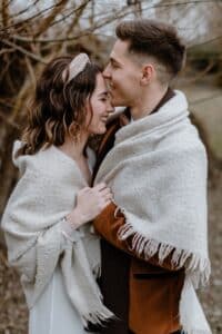 Ein Paar steht eng beieinander, eingehüllt in eine kuschelige Decke mit Fransen. Sie lächeln und schauen einander in die Augen, umgeben von kahlen Ästen. Die Frau trägt ein Stirnband und der Mann eine braune Jacke.