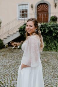 Eine Frau in einem weißen Kleid steht lächelnd auf einem Kopfsteinpflasterweg. Sie hat langes Haar mit einem Blumenschmuck und trägt große Ohrringe. Hinter ihr ist ein Gebäude mit einer Holztür, Fenstern und Grünpflanzen zu sehen.