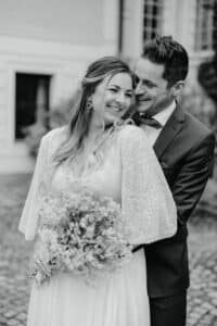 Ein Schwarzweißfoto eines lächelnden Paares im Hochzeitskleid. Die Braut hält einen Blumenstrauß in der Hand und trägt ein Kleid mit strukturierten Ärmeln. Der Bräutigam steht hinter ihr und umarmt sie im Freien in der Nähe eines Gebäudes.