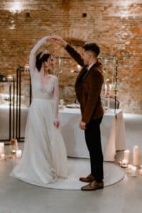 Eine Braut in einem fließenden weißen Kleid und ein Bräutigam in einer braunen Jacke tanzen in einem warm beleuchteten Raum mit unverputzten Ziegelwänden. Kerzen und dekorative Lichter sorgen für eine romantische Atmosphäre.