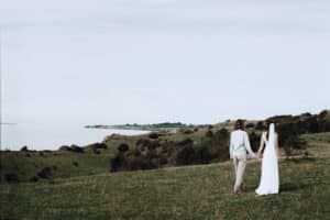 Ein Paar, die Frau trägt ein weißes Kleid und einen Schleier, geht Hand in Hand über einen grasbewachsenen Hügel mit Blick auf eine malerische Küste. Der Himmel ist bedeckt, was der Landschaft eine ruhige und heitere Atmosphäre verleiht.