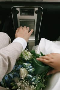 Ein Paar sitzt in einem Auto. Eine Person trägt einen hellbraunen Anzug und bedient die Steuerung des Autos, während die andere Person in einem weißen Outfit einen Strauß blauer und weißer Blumen hält und ihre Hand daneben ruht. Beide zeigen Eheringe.